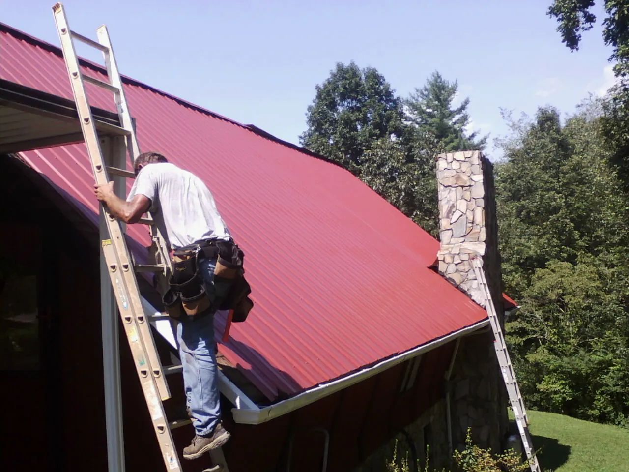 Roofer on a ladder beside a red metal roof and stone chimney — Soffit & Fascia Repair by Roofing Brothers in Grissom Arb